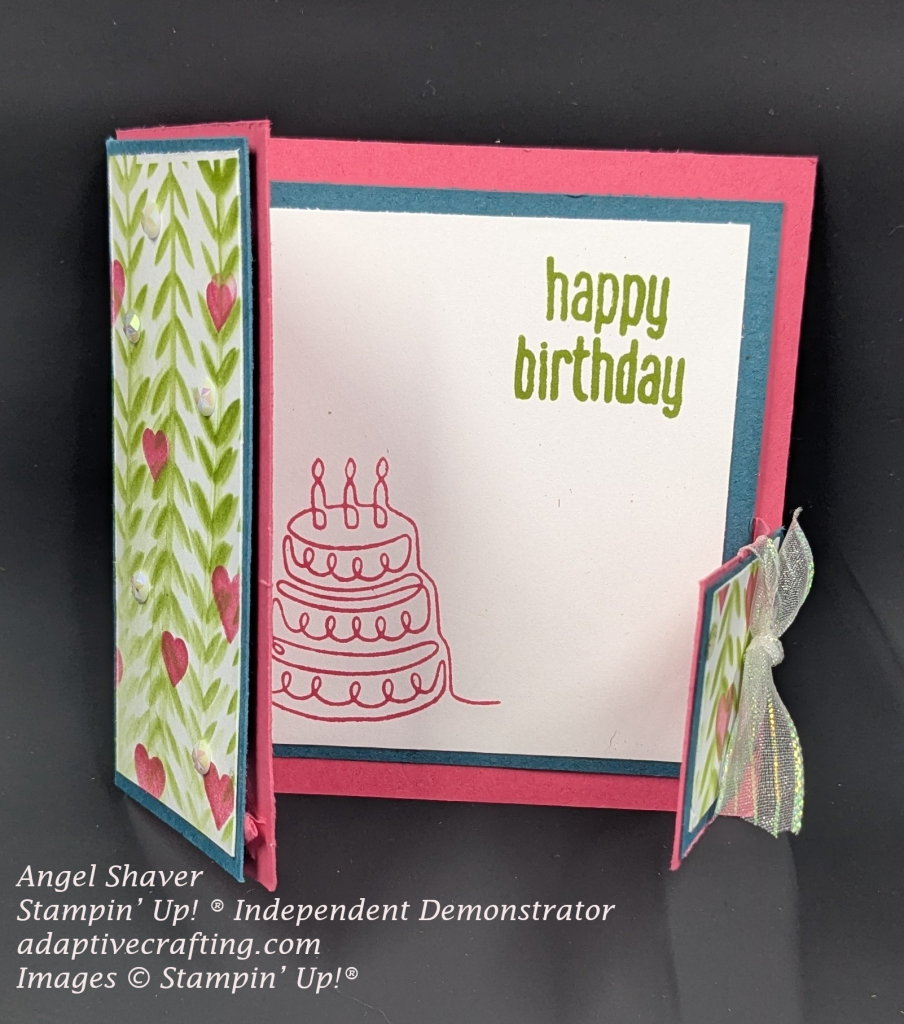 Pink card with blue cardstock framing white rectangles.  Both front rectangle flaps have green leafy vines and pink hearts stenciled on them. The card is beginning to open to reveal the inside of the card.  You can see part of the card with the sentiment "happy birthday." snd a birthday cake.