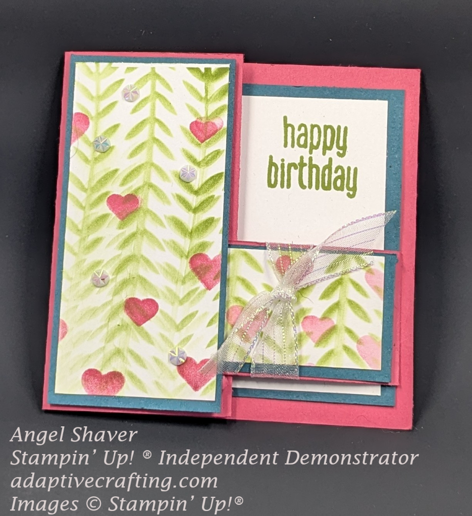 Pink card with blue cardstock framing white rectangles.  Both front rectangle flaps have green leafy vines and pink hearts stenciled on them.  When the card is closed on flap vertically covers the left half of the card and a horizontal flap comes from the right side and tucks into vertical flap.  It is tied with a ribbon.  You can see part of the card interior peeking out with the sentiment "happy birthday."