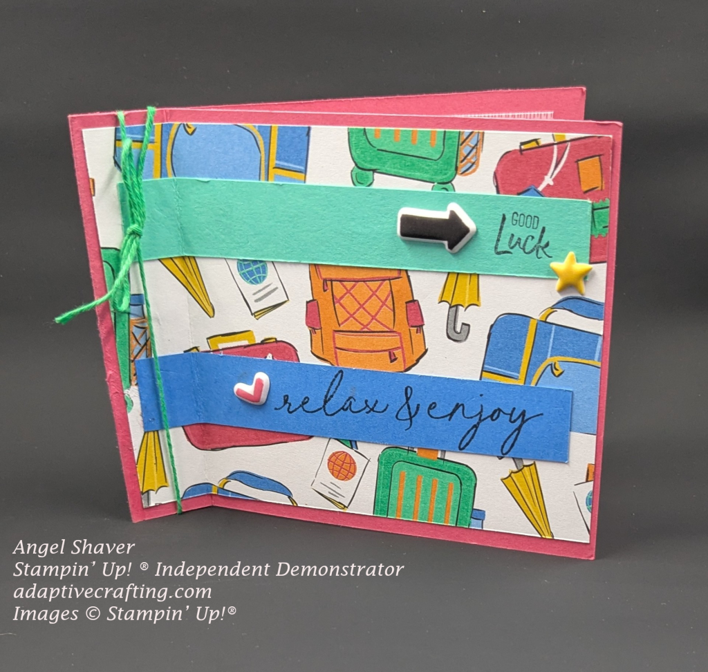 Reddish book fold card with patterned paper showing travel bags, umbrella, passports.  Green twine is tied around the "spine" of the book fold.  Green strip of cardstock is toward the top of the card and says, "good luck" and has a black arrow and yellow stars puffy sticker on it.  There is a strip of blue cardstock toward the bottom of the card that says, "relax & enjoy" and has a red puffy heart sticker.
