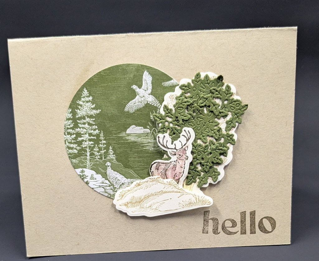 Tan card with green circle of patterned paper with trees and pheasants.  There is a die of a tree with green die cut leaves.  In front of the of the tree is a stamped and cut out deer and rocks.
