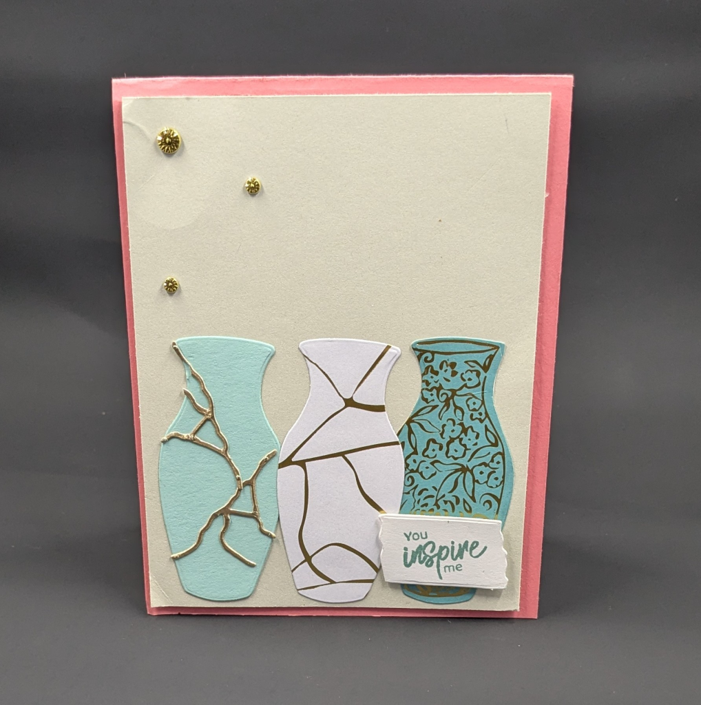 Pink card with beige card front layer.  Three vases are in a row at the bottom of the card.  One is teal with gold flowers and leaves on it.  One is white with gold lines.  One is lighter teal with gold die cut pieces that look like filled cracks in the pottery.  They are supposed to represent kintsugi.
