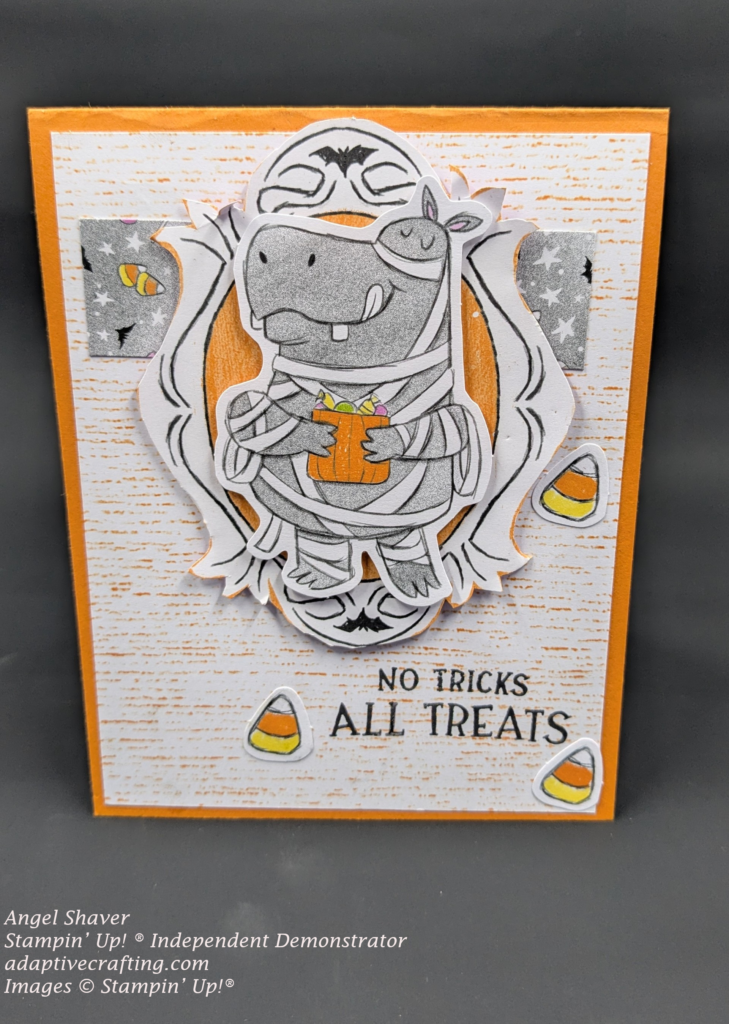 Orange card with patterned background paper that is white with orange stripes.  Strip of gray patterned paper with candy corn, white stars, and bats. Focal image is an orane oval with black curved lines and bats.  On top of the frame is a hippo dressed in white strips like a mummy holding an orange bag full of candy  Three pieces of candy corn are scattered on the card front.  Sentiment says, "No tricks  All treats"