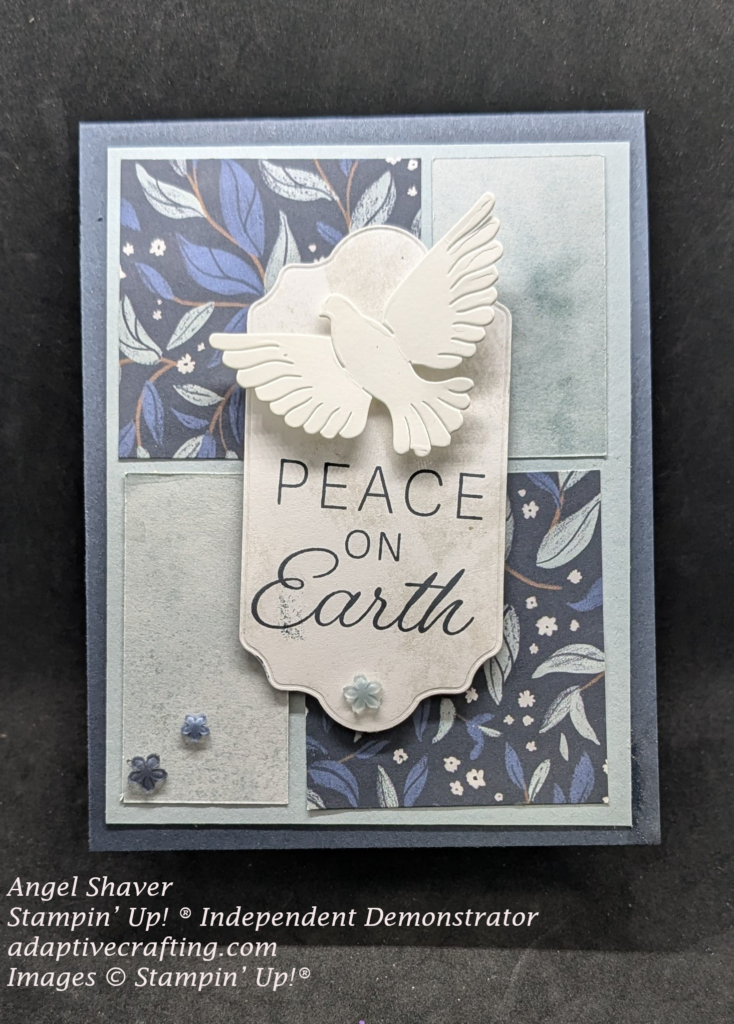 Blue card with background of rectangles of two patterns of background paper. Two are dark blue with lighter blue leaves on stems.  Two are just various shades of light blue.  There is a sentiment label in the center that says, "Peace on Earth" and has a diecut of a flying dove. Light blue flower shaped embellishments are added
