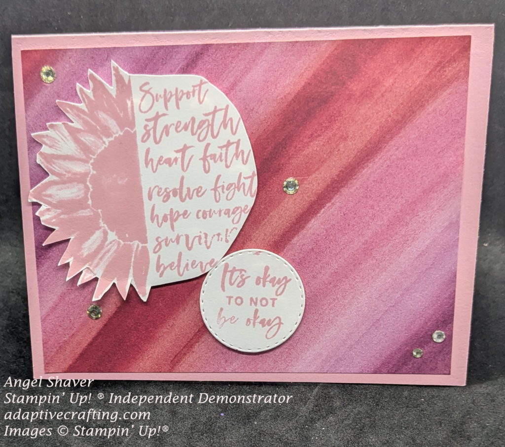 Pink card with pin5 patterned paper diagonal stirpes in different shades of pink.  Stamp 1/2 sunflower with 1/2 circle "support, strength, heart, faith, hope, courage, survivor, believe"  Sentiment label that says, "It's okay to not be okay."  Decorated with rhinestones.