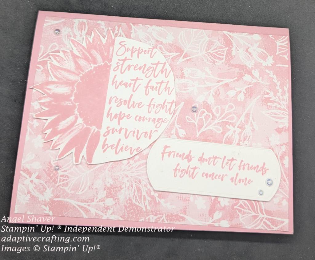 Pink card with pin patterned paper with white leaves.  Stamp 1/2 sunflower with 1/2 circle "support, strength, heart, faith, hope, courage, survivor, believe"  Sentiment label that says, "Friends don't let friends fight cancer alone."  Decorated with iridescent pearls.