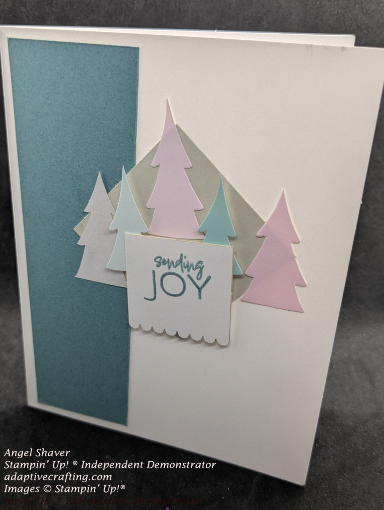 White card with teal cardstock strip on left side of paper   Light brown diamond shape in center of card with tree dies in shades of pink, teal, and white.  Sentiment label with scallops across bottom says, "sending joy."