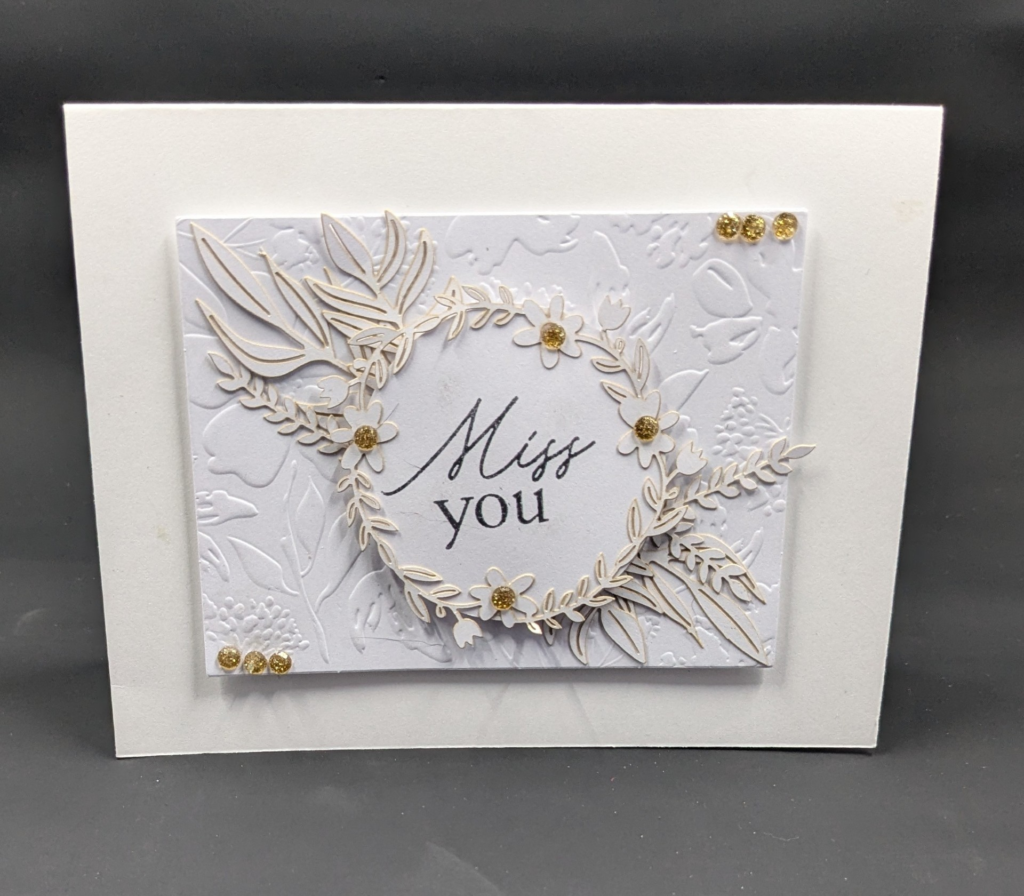 White on white card.  White card with white embossed layer a focal point.  Ephermera leaves going diagonally from corner to corner with sentiment circle in middle that says, "Miss you" and is surrounded by a floral wreath.  Gold embellishments are added in corners and centers of flowers.