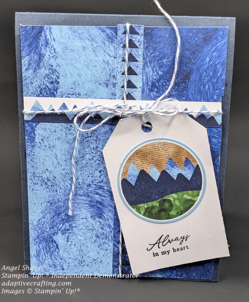 Blue card with blue patterned paper layer with various shades/swirls of diferent blue colors.  Printed sticked with blue triangle goes vertically up center of card.  Printed stick with mountains runs horizontally.  Blue and white twine is strung across both stickers and tied in a bow where they meet.  White tag is tied to the twine.   TAg has a circle sticker on it with mountain landscape and sentiment that says, "Always in my heart."
