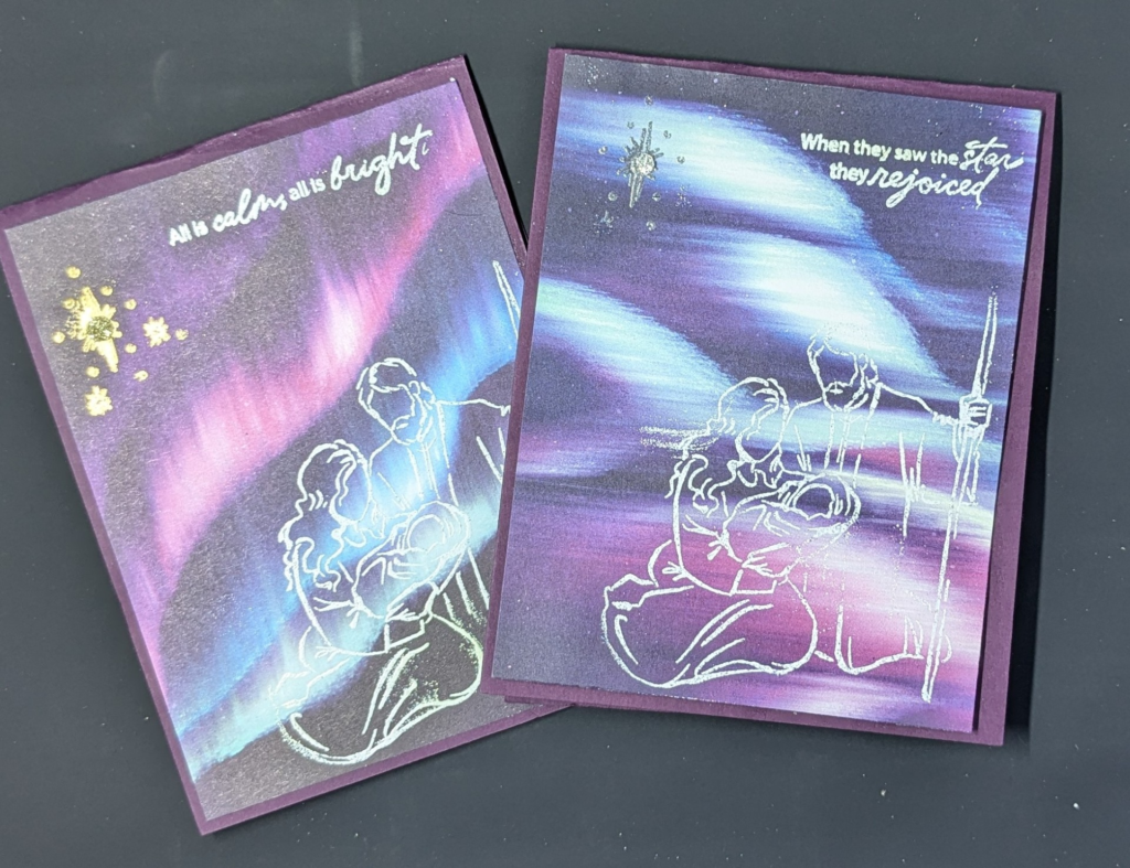 Two purple cards with patterned paper layers that look like the lights of aurora in purple, pink, and white. Holy family and sentiment in heat embossed in white. Star is heat embossed in silver on one and gold on the other.