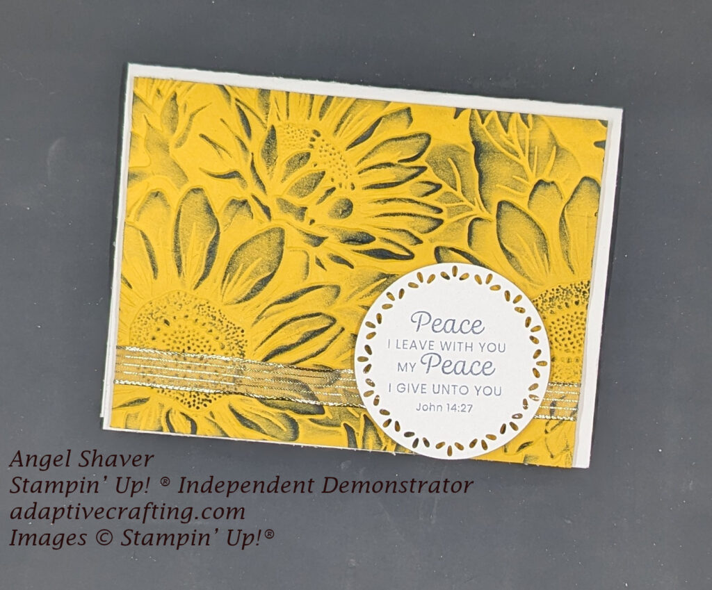 White card with yellow card front embossed with sunflowers with are then highlighted with navy ink. Mesh ribbon with gold stripes runs across bottom front of card. There is a white sentiment label circle image that says, "Peace I leave with you, my peace I give unto you. John 14:27"