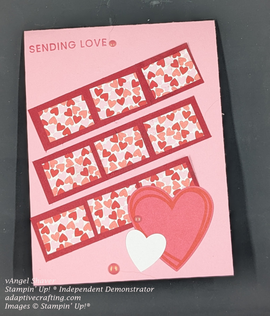 Pink card with three strips of red cardstock going diagonally across front of card with three rectangles of patterned paper with pink and red hearts in each one.  Sticker of large red heart and small white heart is at the bottom of the page.  Sentiment "sending love" is stamped at the top and card is decorated with red enamel dots
