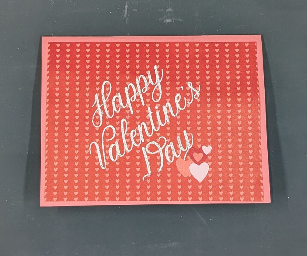 Pink card with red patterned paper with pink hearts. "Happy Valentine's Day" is heat embossed in white. Red, white, and pink heart stickers are added for embellishments.