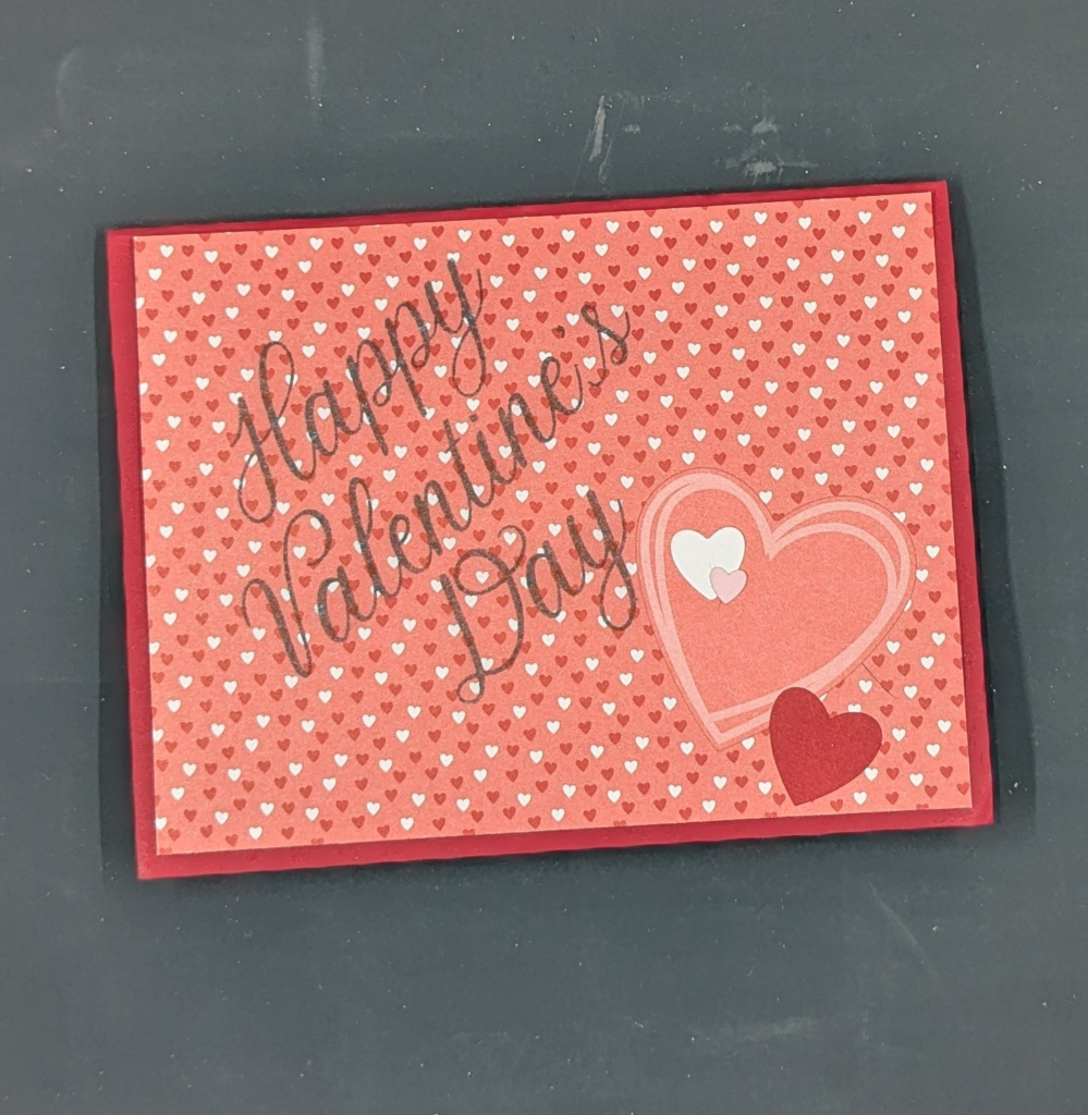 Red card with pink patterned paper with red and white hearts. "Happy Valentine's Day" is stamped in black. Red, white, and pink heart stickers are added for embellishments.