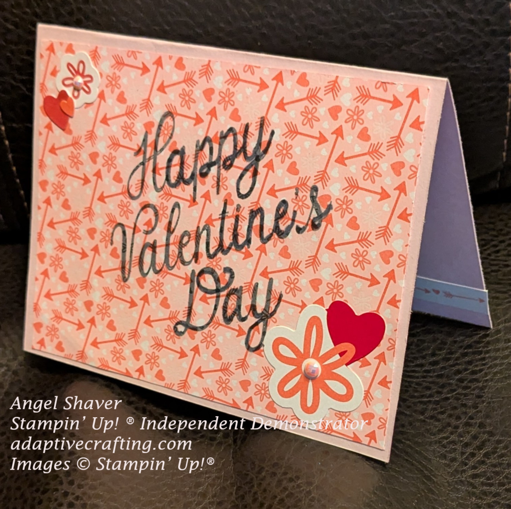 Pink card with layer of printed paper that is pink with printed hearts, flowers, and arrows. "Happy Valentine's Day" stamped in black in center of card. Stickers of flowers and hearts two corners. The flowers have faceted gems in the center