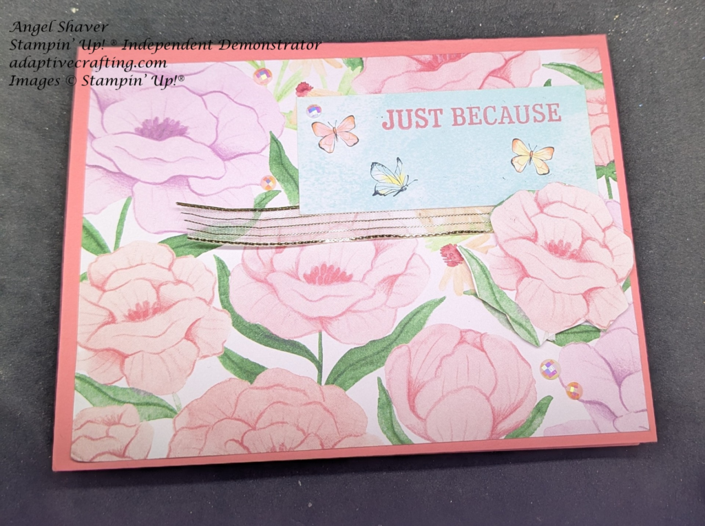 Pink card with layer of patterned paper with pink and purple flowers on front.  Rectangle with butterflies flying through blue sky and "Just Because" stamped in pink is on top right of card front.  Gold striped ribbon is bordering the bottom of the card with a large pink flower at the bottom right of sentiment label.  Varying shades of pink faceted gems are on the card.