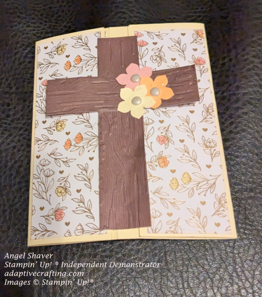 Yellow card with strips of patterned paper featuring foil outlined flowers and hearts. Some of the flowers were colored in with pink, yellow, and peach markers.  The focal point image is a cross made of two strips of woodtone paper embossed with a Birch wood embossing folder.  Three punched flowers--pink, yellow, and peach are added to the front of the cross with vintage pearl centers.