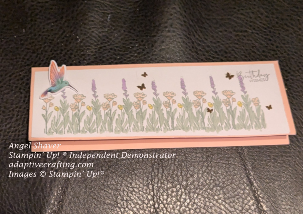 Pink slimline card with white front layer stamped with row of flowers on front to make a flower garden. Flowers are colored pink, purple, and yellow. Sentiment stamped in top right corner says "Birthday wishes" Hummingbird ephemera and butterfly embelishments are added amongst the flowers.