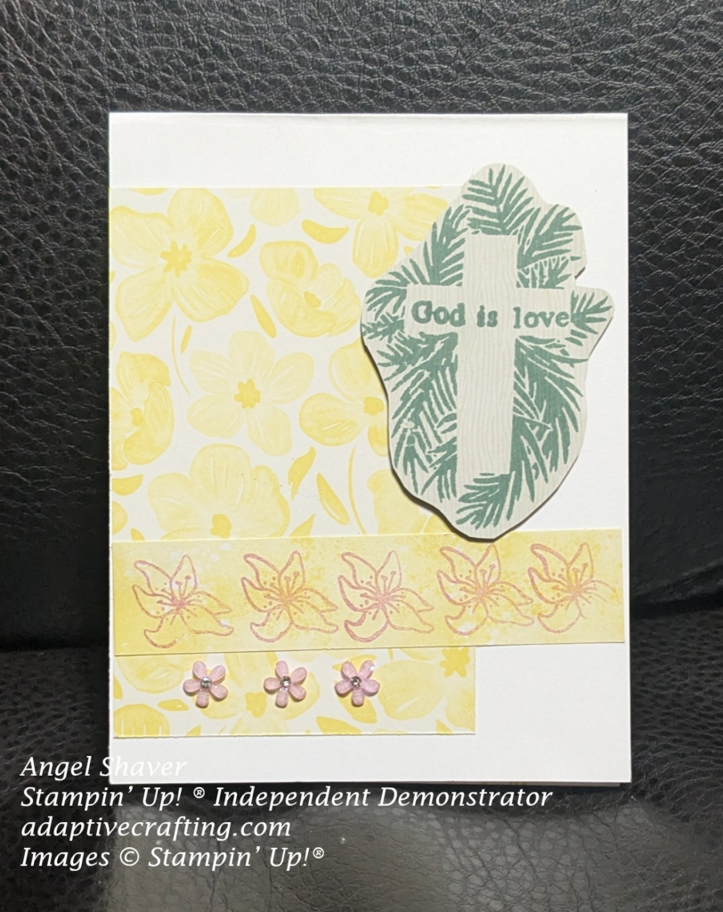 White card with rectangle of patterned paper with yellow flowers There is a strip of yellow paper with stamped purple lilies toward the bottom of the yellow flowers patterned paper.  Focal image is aa wood pattern cross on bed of olive branches.  Sentiment stamped on the cross says, "God is love."  Purple flower embellishments are at the bottom of the card.