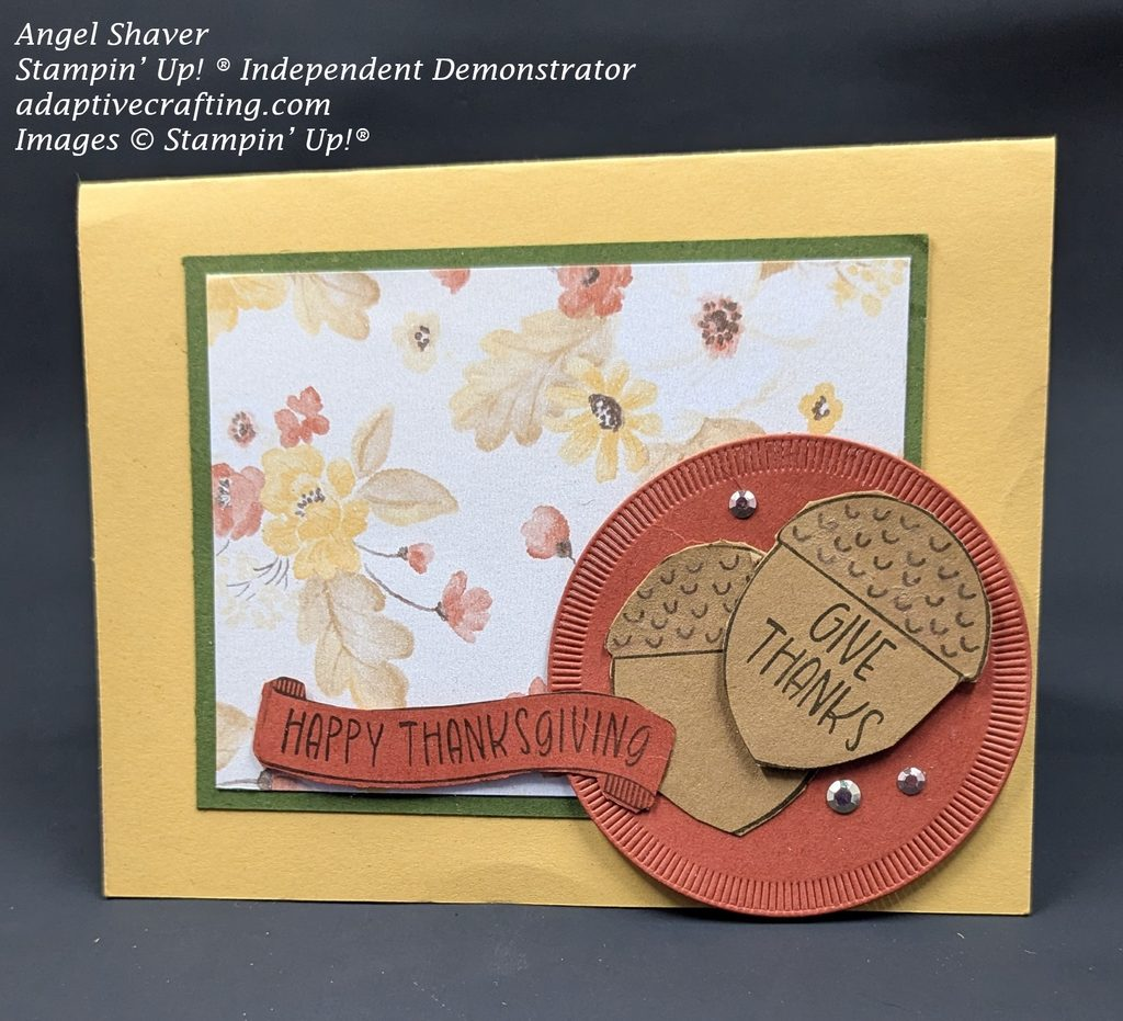 Golden yellow card with patterned paper of fall colored floral arrangements framed with a piece of green cardstock as the focal image.  There is a rust orange circle with two brown acorns on it.  The top acorn says "Give Thanks."  A rust orange sentiment banner says "Happy Thanksgiving."